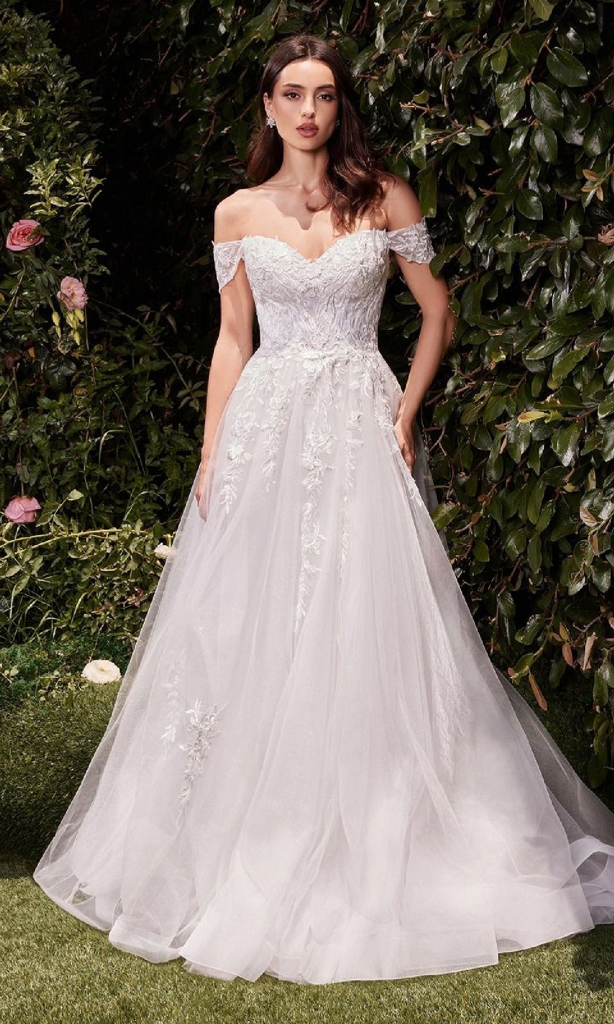 A woman stands outside before green foliage, wearing the Off-White Long Wedding Dress: Ladivine C159W—an elegant off-the-shoulder bridal gown with delicate lace details.